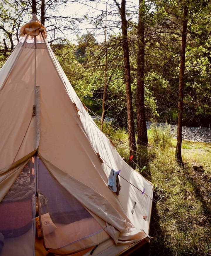 Photo of a beige tipi tent with a half-open entrance, offering a glimpse inside; in the background, a river flows between trees.